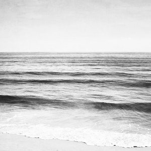 Puede incluir: Fotograf&iacute;a en blanco y negro del oc&eacute;ano. La imagen captura el mar con suaves olas que se acercan a una playa arenosa. El cielo tiene un tono suave y apagado, creando una escena costera serena y minimalista.
