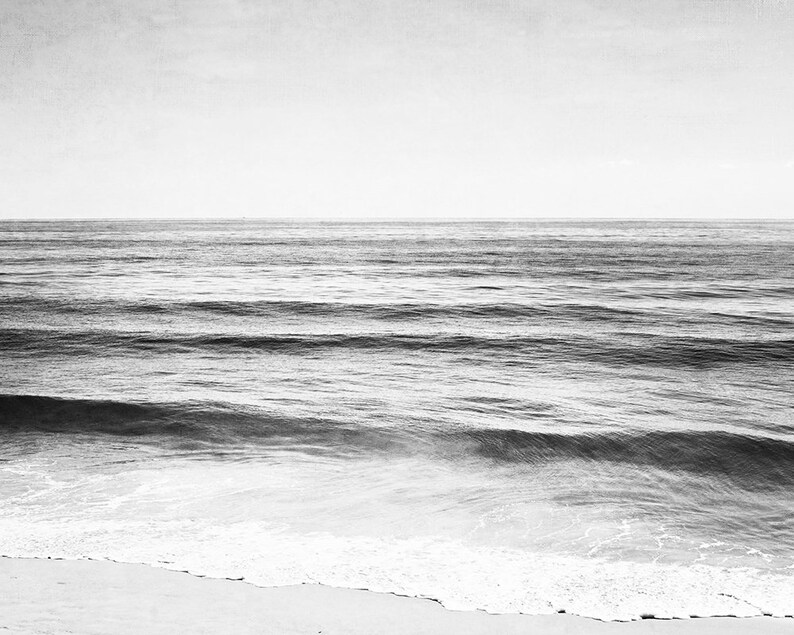 Puede incluir: Fotograf&iacute;a en blanco y negro del oc&eacute;ano. La imagen captura el mar con suaves olas que se acercan a una playa arenosa. El cielo tiene un tono suave y apagado, creando una escena costera serena y minimalista.
