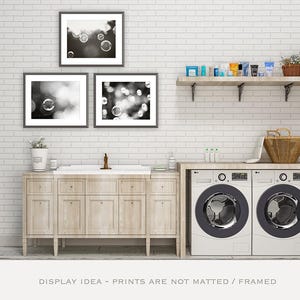 May include: A laundry room with a white brick wall, featuring framed black and white bubble art. A wooden shelf holds toiletries, and a countertop supports a washing machine and dryer. A wooden cabinet and a basket complete the scene.