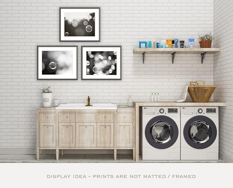 May include: A laundry room with a white brick wall, featuring framed black and white bubble art. A wooden shelf holds toiletries, and a countertop supports a washing machine and dryer. A wooden cabinet and a basket complete the scene.