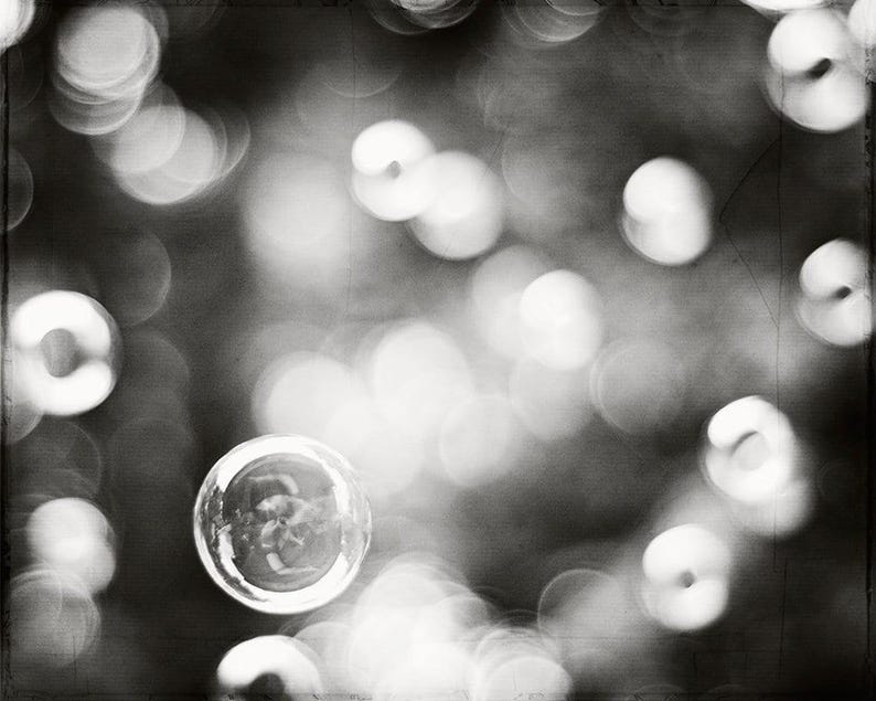 May include: Monochrome photograph with a shallow depth of field, showcasing blurred circular lights. A single, transparent bubble is in focus, reflecting its surroundings. The image has a vintage, grainy texture.