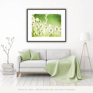 Pode incluir: Uma impress&atilde;o de arte emoldurada com flores silvestres brancas em um fundo verde suave. A impress&atilde;o &eacute; exibida acima de um sof&aacute; cinza com almofadas verdes e um cobertor correspondente. Uma lumin&aacute;ria de ch&atilde;o e uma planta decorativa completam o ambiente.