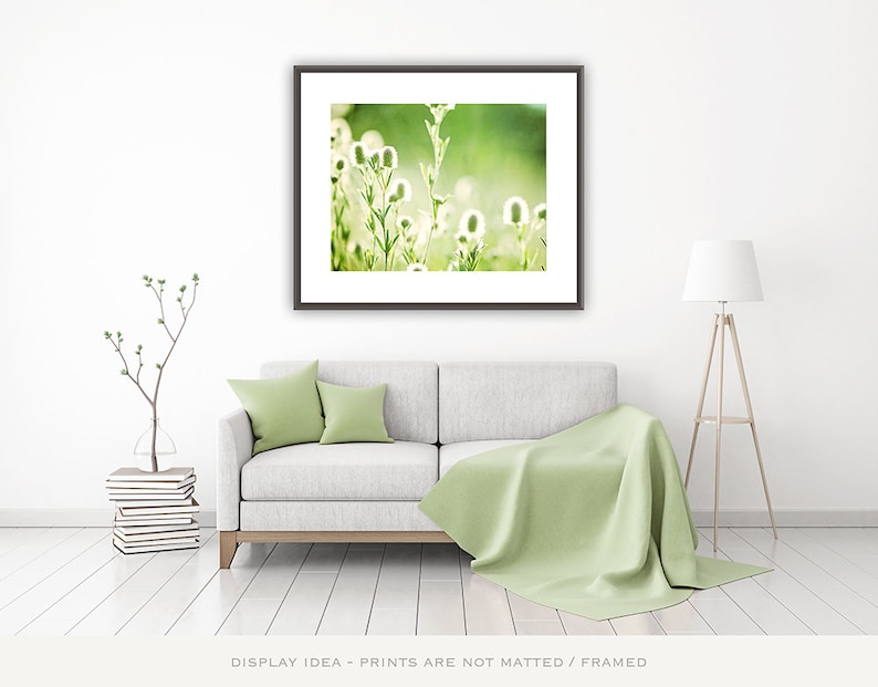 Pode incluir: Uma impress&atilde;o de arte emoldurada com flores silvestres brancas em um fundo verde suave. A impress&atilde;o &eacute; exibida acima de um sof&aacute; cinza com almofadas verdes e um cobertor correspondente. Uma lumin&aacute;ria de ch&atilde;o e uma planta decorativa completam o ambiente.