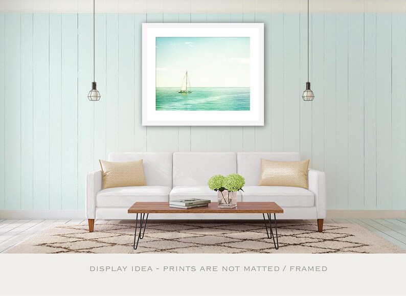 May include: A framed print of a sailboat on the ocean hangs above a white sofa with yellow pillows. A wooden coffee table with black hairpin legs sits on a patterned rug. The wall is painted a light blue.