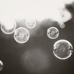 May include: Monochrome image of multiple soap bubbles floating in the air. The bubbles are spherical and clear, with reflections of the surroundings visible. The background is blurred, with bright light shining through.