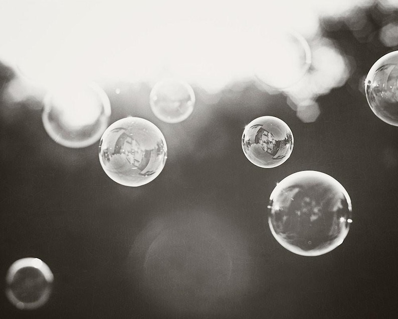May include: Monochrome image of multiple soap bubbles floating in the air. The bubbles are spherical and clear, with reflections of the surroundings visible. The background is blurred, with bright light shining through.