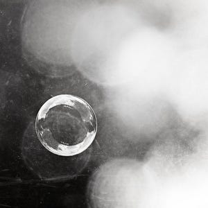May include: A monochrome image featuring a clear, spherical bubble in sharp focus. The bubble reflects its surroundings. The background is blurred with soft, out-of-focus highlights, creating a dreamy effect. The image has high contrast and a minimalist aesthetic.