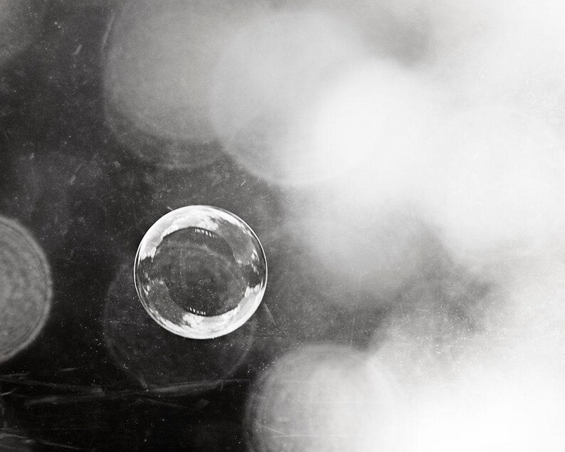 May include: A monochrome image featuring a clear, spherical bubble in sharp focus. The bubble reflects its surroundings. The background is blurred with soft, out-of-focus highlights, creating a dreamy effect. The image has high contrast and a minimalist aesthetic.
