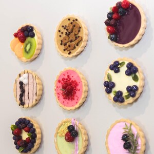 May include: Nine miniature tarts with diverse toppings, including fruit and chocolate. The tarts have a golden-brown, fluted crust. Toppings include blueberries, raspberries, kiwi, and chocolate chips. The tarts are arranged on a white surface.
