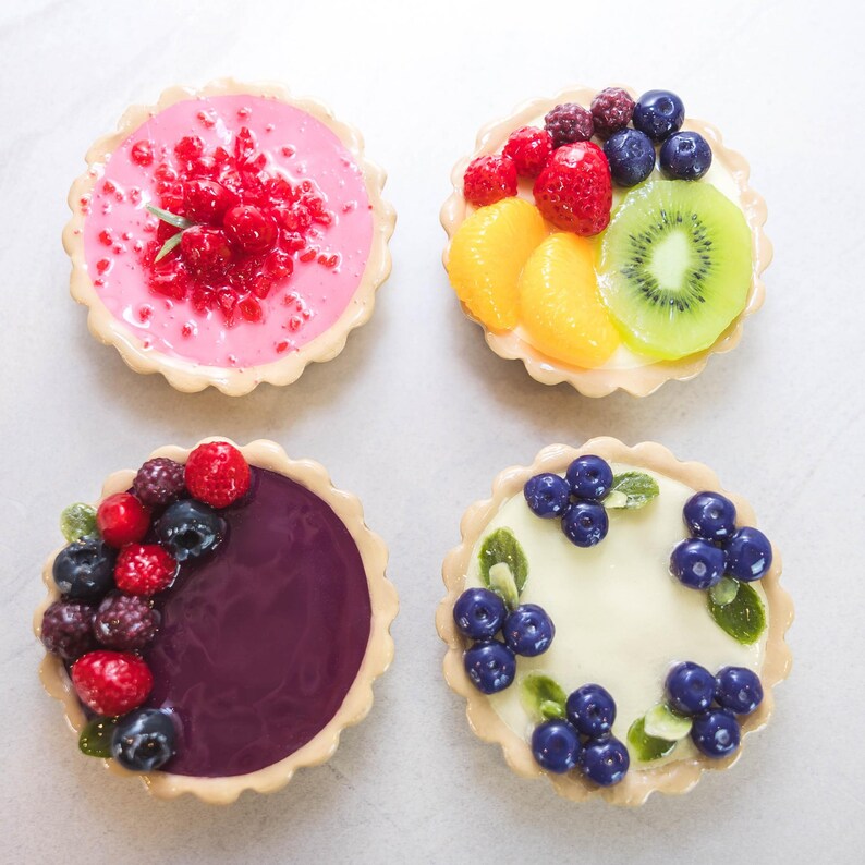 May include: Four miniature fruit tarts with a light-coloured, scalloped crust. Each tart is topped with a different arrangement of colourful fruit, including raspberries, blueberries, kiwi, and mandarin orange slices. The fillings are pink, white, and purple.
