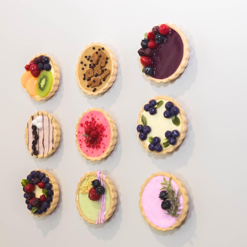 May include: Nine miniature tarts with diverse toppings, including fruit and chocolate. The tarts have a golden-brown, fluted crust. Toppings include blueberries, raspberries, kiwi, and chocolate chips. The tarts are arranged on a white surface.
