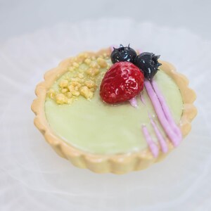 May include: A miniature tart with a pale green filling, decorated with a red strawberry, two dark blueberries, and golden crumbs. The tart has a fluted, light brown crust and pink drizzle.