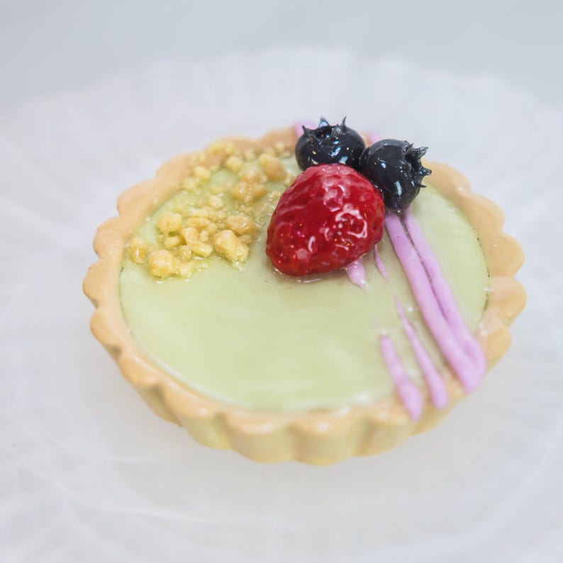 May include: A miniature tart with a pale green filling, decorated with a red strawberry, two dark blueberries, and golden crumbs. The tart has a fluted, light brown crust and pink drizzle.