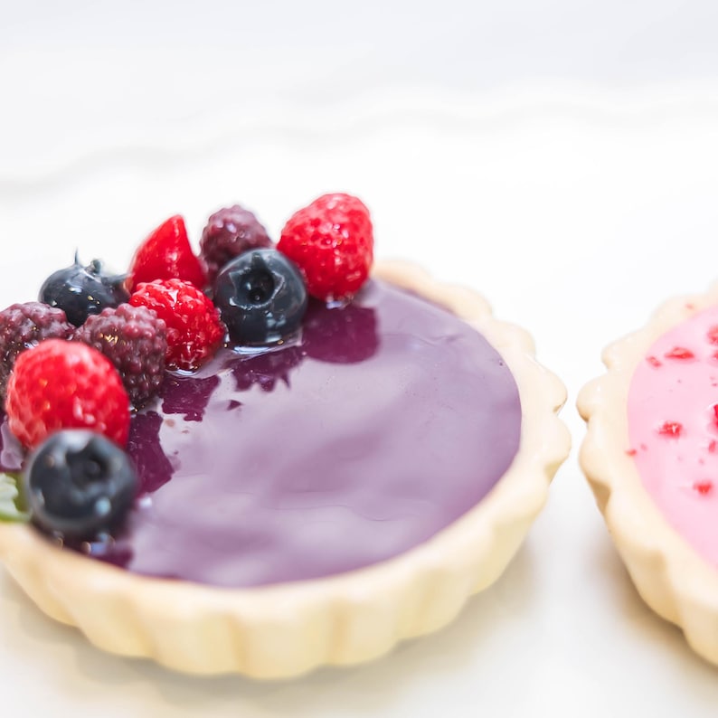 May include: Two mini tarts are shown, one with a purple glaze and fresh berries: blueberries, raspberries, and blackberries. The other has a pink glaze with red sprinkles. Both have a light-coloured, fluted pastry crust.