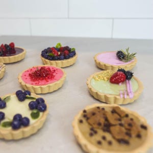 May include: A selection of miniature tarts with diverse toppings, such as berries and chocolate chips. The tarts feature a light-coloured, fluted pastry crust and are arranged on a light-coloured surface.