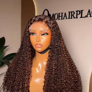 May include: A brown curly wig displayed on a mannequin head. The wig features a lace front and is styled with tight curls. The mannequin is mounted on a tripod. The text "MOHAIRPLACE" is visible in the background.