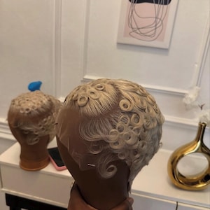 May include: A blonde wig with finger waves is displayed on a mannequin head. The wig has a lace front and is styled with tight, rounded curls. Another wig is visible in the background. The wig is likely for cosplay or fashion.