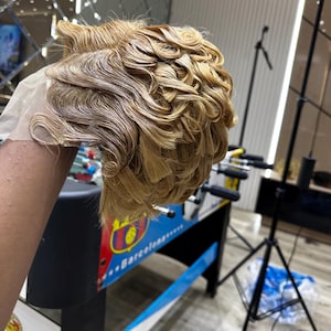 Puede incluir: Peluca castaño claro con cabello corto y ondulado. La peluca tiene una parte delantera de encaje y está peinada con una raya a un lado. El cabello es de un color dorado cálido y parece recién peinado. La peluca es probablemente para moda o cosplay.