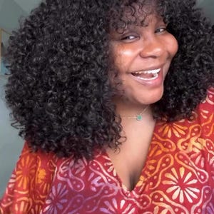 May include: A person with dark, curly hair smiles, wearing a red and orange patterned top. The top has a V-neck and a floral design. A gold necklace with a small pendant is visible.