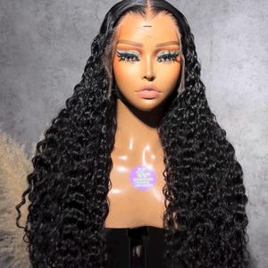 May include: A mannequin head displaying a long, black, curly wig. The wig features a lace front and is styled with voluminous, cascading curls. The mannequin has makeup applied, including eyeshadow and long eyelashes. The background is a solid gray.