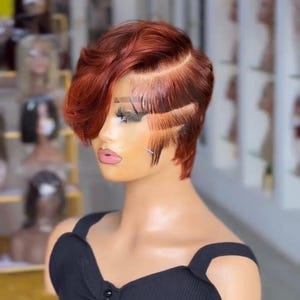 May include: A mannequin head displaying a short, auburn-colored wig with a side-swept bang and intricate side detailing. The mannequin is wearing a black, ribbed top with button accents. The background is a blurred display of other wigs.