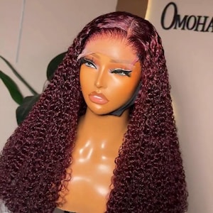 May include: A burgundy curly wig displayed on a mannequin head. The wig has a lace front and is set against a neutral backdrop. The mannequin is mounted on a tripod.