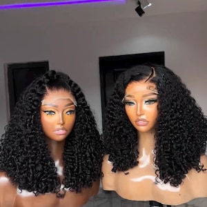 May include: Two mannequin heads with curly black wigs. The wigs have a natural hairline and are styled with loose curls. The mannequins are on tripods, and the background is a neutral color.