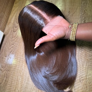 May include: A dark brown wig with a centre parting. The wig is styled with long, straight hair. The hair is shiny and appears to be in good condition. The wig is being held by a hand.
