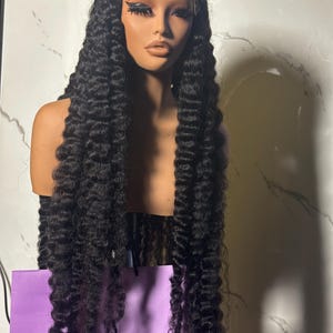 May include: A long, black curly wig displayed on a mannequin head. The wig has a lace front and is styled with voluminous, tight curls. The mannequin has makeup applied, including dark eyelashes and lipstick. A purple bag with the text "Sussy Hairworks" is visible.