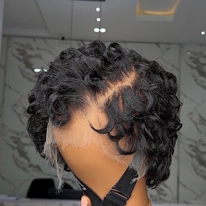 Könnte beinhalten: Kurze, lockige schwarze Perücke auf einem Schaufensterkopf. Die Perücke hat eine Spitze vorne und verstellbare Träger. Das Haar ist in engen, voluminösen Locken gestylt und bietet einen natürlichen Look. Der Schaufensterkopf ist hellbraun.