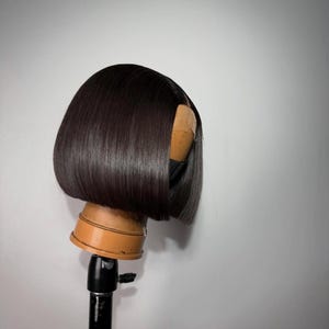 Può includere: Una parrucca a caschetto color marrone scuro, liscia e lucente, con un taglio netto e dritto. I capelli presentano una riga centrale che rivela la base in pizzo. La parrucca è esposta su una testa di manichino beige su un supporto nero.