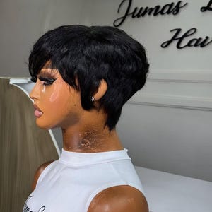 Puede incluir: Una peluca negra de corte pixie en un maniquí. La peluca tiene el pelo corto y en capas con un flequillo lateral. El maniquí lleva una camiseta sin mangas blanca con el texto "Jumas Hair". El fondo es una pared blanca con el texto "Jumas Hair" en cursiva.