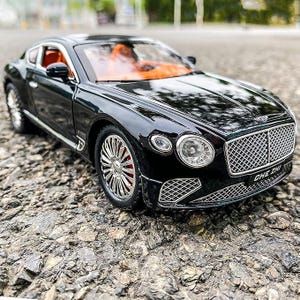 May include: A black toy car with silver wheels and red brake callipers. The car features a detailed front grille and headlights. The interior is orange. The car is on a rocky surface. The text "CHE ZHI" is visible on the front.