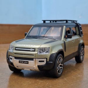 Puede incluir: Modelo a escala de un SUV dorado con detalles en negro, incluyendo baca y neumáticos negros. El texto "CHE ZHI" aparece en la matrícula. La palabra "DEFENDER" es visible en el capó.