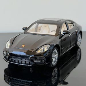 May include: A black Porsche Panamera scale model car featuring a glossy exterior finish, silver multi-spoke wheels, and a light beige interior visible through the windows. The front hood displays the Porsche emblem in red, black, and gold.