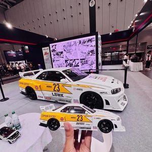 May include: A white and yellow model car with the number 23 and the text "LBWK" and "LIBERTY WALK". A larger version of the same car is behind it. The cars are on display at an event.