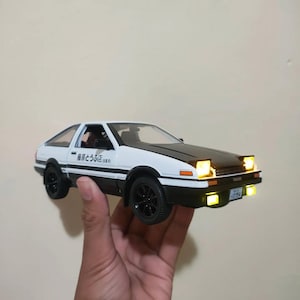 May include: A white and black scale model car with a black bonnet, pop-up headlights illuminated in yellow, and yellow fog lights. The side features Japanese text that reads Fujiwara Tofu Ten. The car has black wheels and a front number plate.