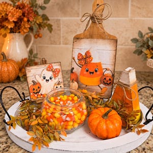 May include: A white round tray with black handles holds a glass bowl filled with candy corn, a small orange pumpkin, a wooden cutting board with a candy corn character illustration, a square sign with a ghost illustration, and faux foliage.