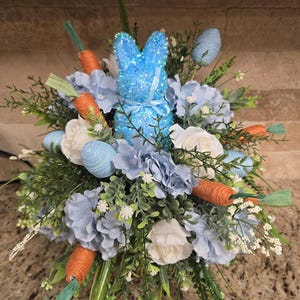 May include: A festive Easter arrangement featuring a sparkling blue bunny, surrounded by light blue and white flowers, faux carrots, and blue decorative eggs. The arrangement is set against a neutral background, creating a cheerful spring display.