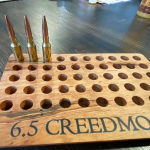 May include: A wooden ammunition holder with multiple round holes, holding three bullets. The holder is engraved with the text "6.5 CREEDMOOR". The bullets have a brass casing and a pointed tip.