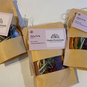 May include: Three brown paper bags with clear windows, each containing a mix of dried herbs and spices. Attached are pink business cards with the "Hanley Homemade" logo and contact information.