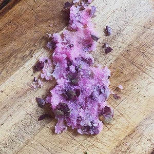 May include: A close-up shot of a pile of pink, granulated sugar mixed with small, dark purple leaves, resting on a wooden cutting board. The sugar appears to be a bath or body scrub, with a textured, crystalline appearance.