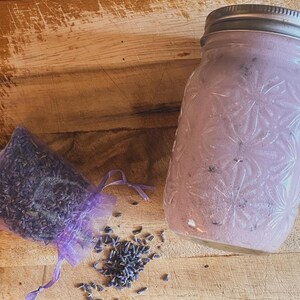 Peut inclure: Un bocal en verre avec un couvercle argenté rempli d'une substance rose-violet, à côté d'un petit sac en filet violet et de boutons de lavande éparpillés sur une surface en bois. Le bocal a un motif floral décoratif.