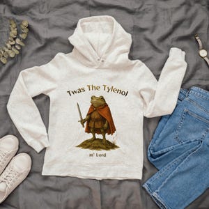 May include: A light gray hoodie featuring a whimsical illustration of a frog dressed as a medieval knight, holding a sword and wearing a red cape. The text "Twas The Tylenol m' Lord" is printed above and below the frog graphic.