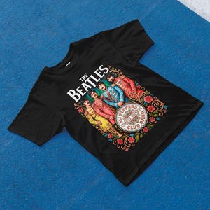 May include: Black t-shirt with a vibrant graphic of The Beatles. The design showcases the band's name, the album title "Sgt. Pepper's Lonely Hearts Club Band," and illustrations of the band members in their famous attire. The graphic is framed by floral patterns.