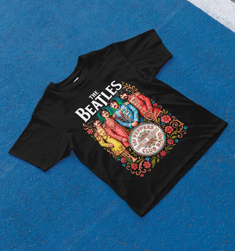 May include: Black t-shirt with a vibrant graphic of The Beatles. The design showcases the band's name, the album title "Sgt. Pepper's Lonely Hearts Club Band," and illustrations of the band members in their famous attire. The graphic is framed by floral patterns.