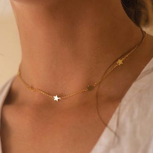 Puede incluir: Un delicado collar dorado con pequeños dijes en forma de estrella. El collar se lleva alrededor del cuello y descansa sobre la piel. Las estrellas están espaciadas uniformemente a lo largo de la cadena, creando un diseño simple pero elegante.