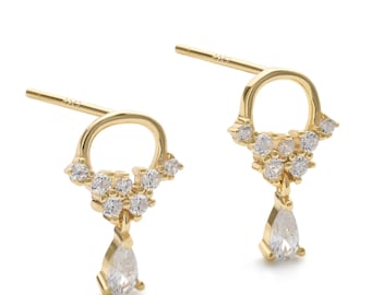 May include: A pair of gold-coloured stud earrings. Each earring has a small, open circle design with a cluster of small, clear gemstones. A pear-shaped gemstone dangles from the bottom of each earring. The earrings are set against a white background.