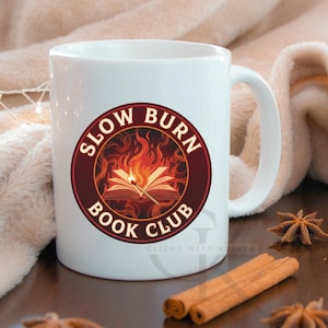 Può includere: Tazza in ceramica bianca con logo "Slow Burn Book Club". Il logo presenta un libro aperto con fiamme e un fiammifero acceso, all'interno di un cerchio bordeaux. La tazza ha un manico curvo ed è appoggiata su una superficie di legno con bastoncini di cannella e anice stellato.