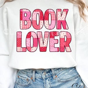 May include: White sweatshirt with the words "BOOK LOVER" in a patchwork design of pink and red patterns. The sweatshirt is paired with blue denim jeans. The overall style is casual and comfortable.